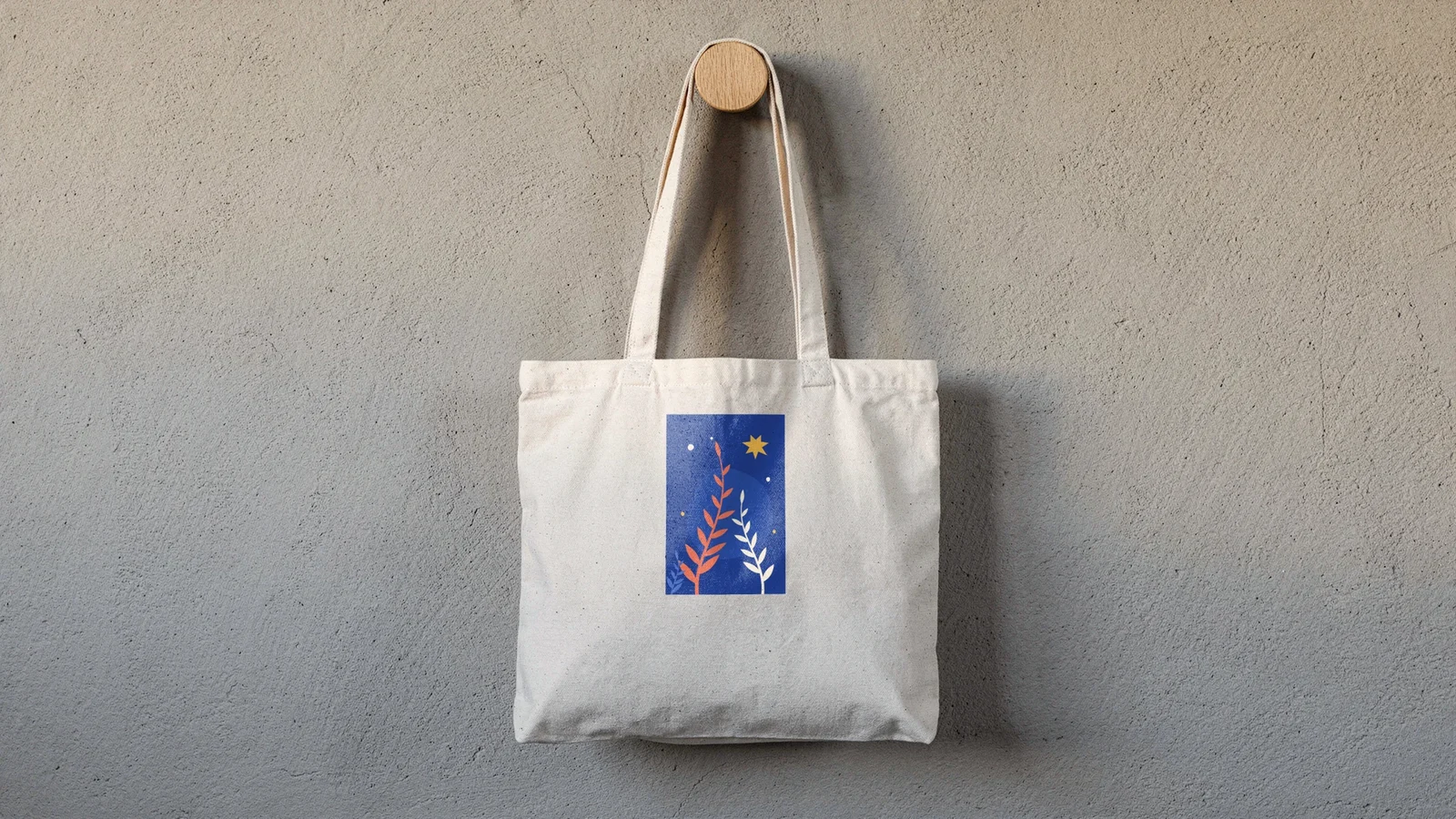 A natural canvas tote bag hanging on a wooden wall peg with a shopper's botanical design on a deep blue panel printed on its front, the canvas weave visible through the print, rendered by SudoMock from a product photo