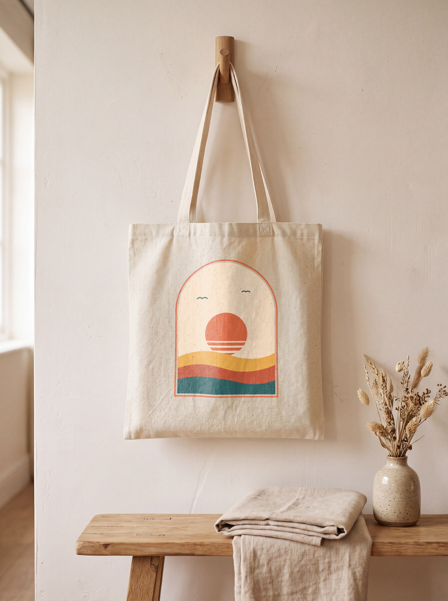 A cream canvas tote bag with a printed sunset arch design hanging from a wooden peg above a bench, a SudoMock render at 1500 by 2010 pixels