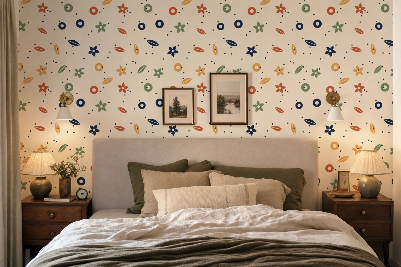 A cream wallpaper with small flowers, leaves and rings covering the wall behind a bed, with two framed prints, two brass sconces and two table lamps in front of the pattern